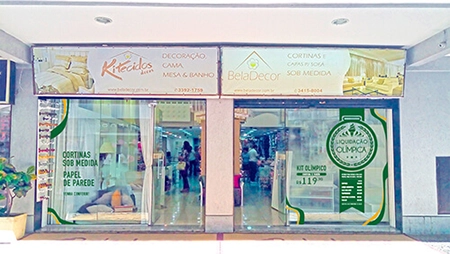 imagem da fachada da loja, com adesivação da promoção liquidação olímpica nos vidros, na cor verde, com a logo da promoção simulando uma medalha e alguns preços das ofertas