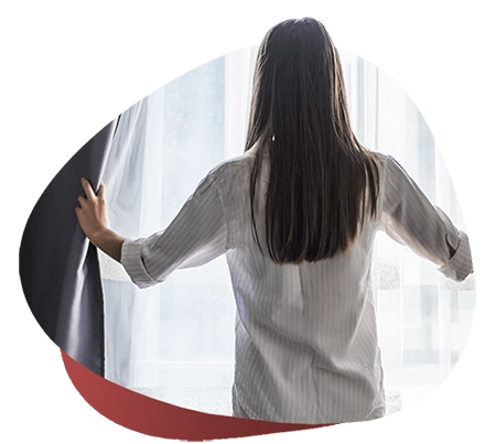 imagem de uma mulher, de cabelo castanho, de costas, com uma camisa social branca listrada em cinza, abrindo a cortina da janela e entrando a luz, sobre um fundo vermelho degradê